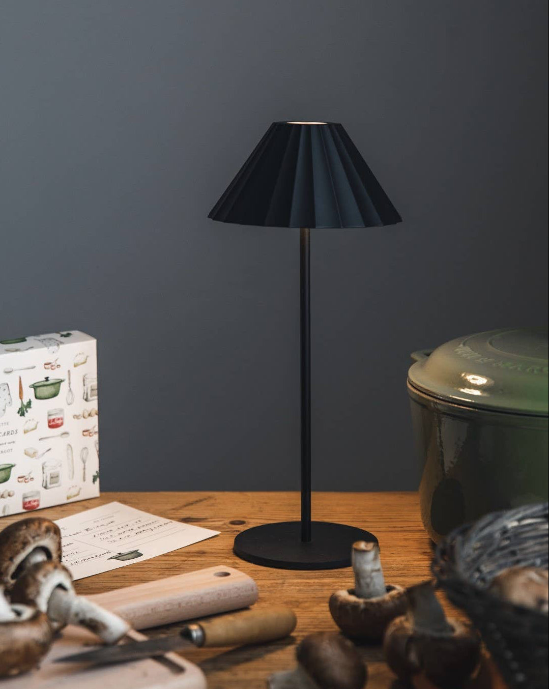 A black cordless bistro lamp with a ruffled shade is displayed on a wooden table with a cutting board and mushrooms around it. The lamp is off, and there is a USB cord visible.
