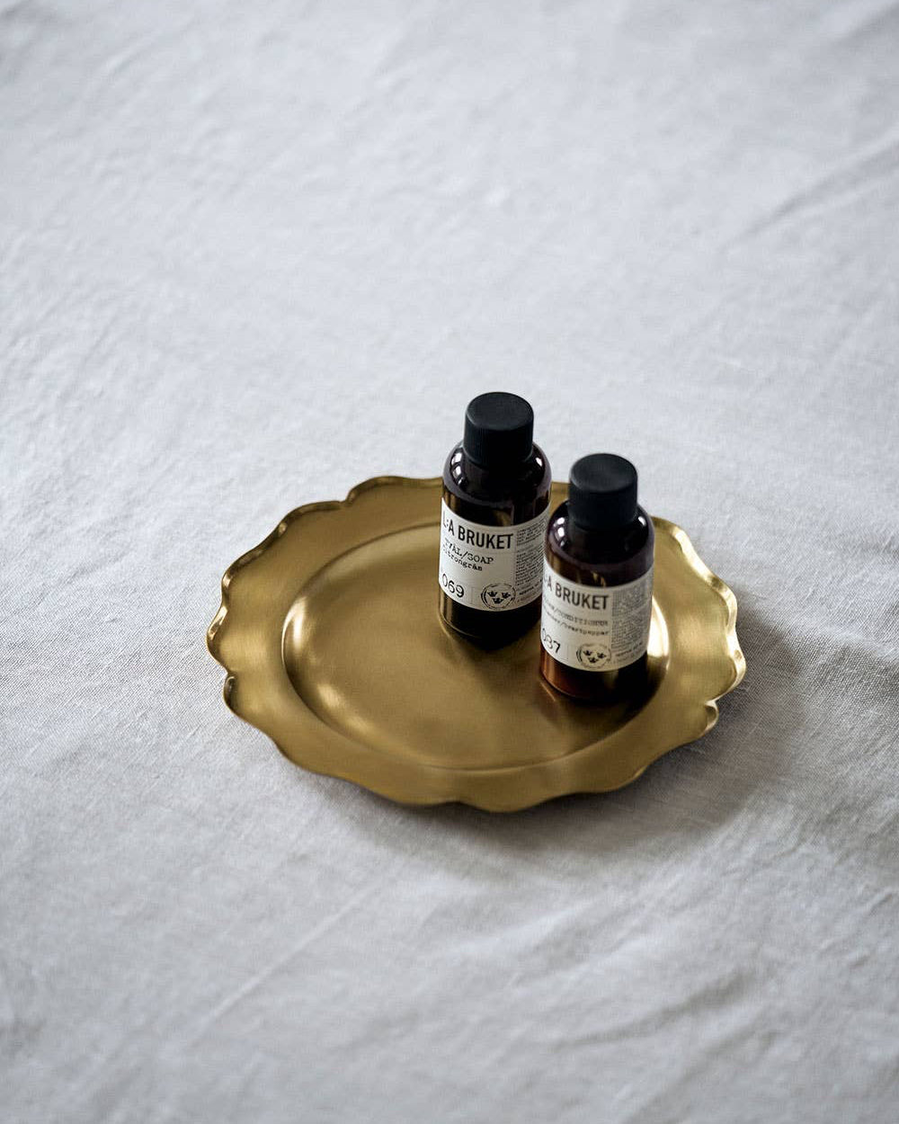Two small bottles with black caps on a gold tray over a white fabric background
