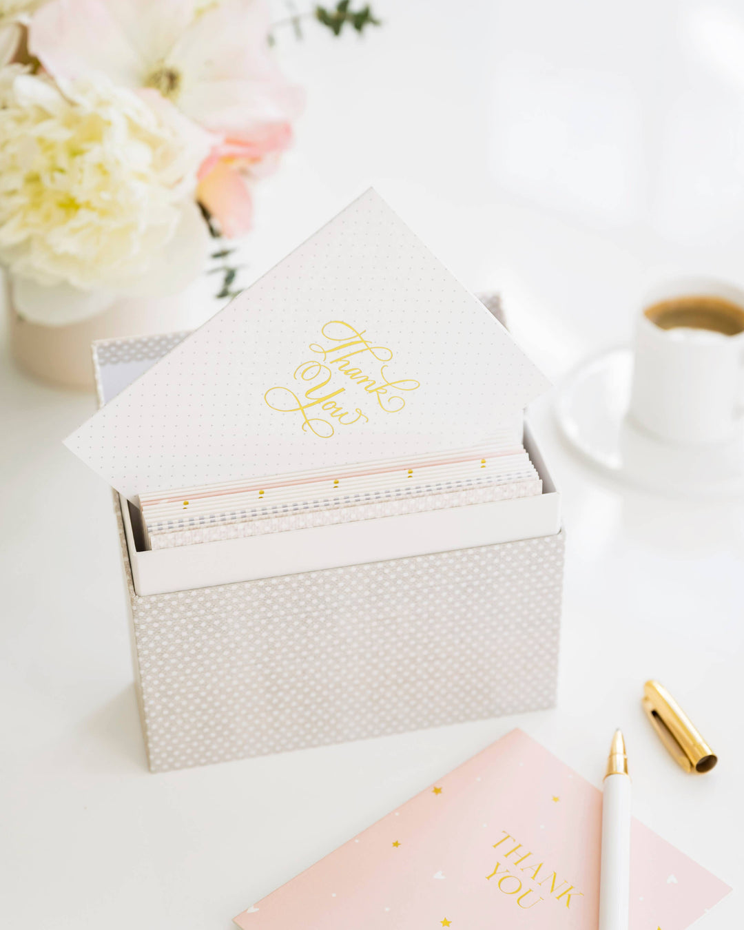 Stationery items including a pink notebook, gold pen, and white box on a light surface with flowers in the background.