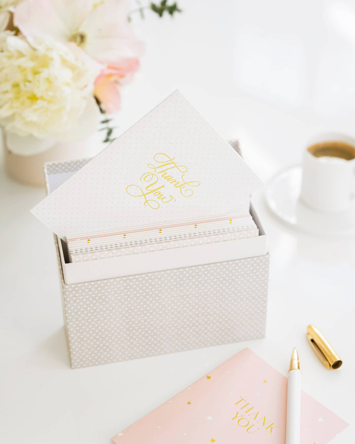 Stationery items including a pink notebook, gold pen, and white box on a light surface with flowers in the background.