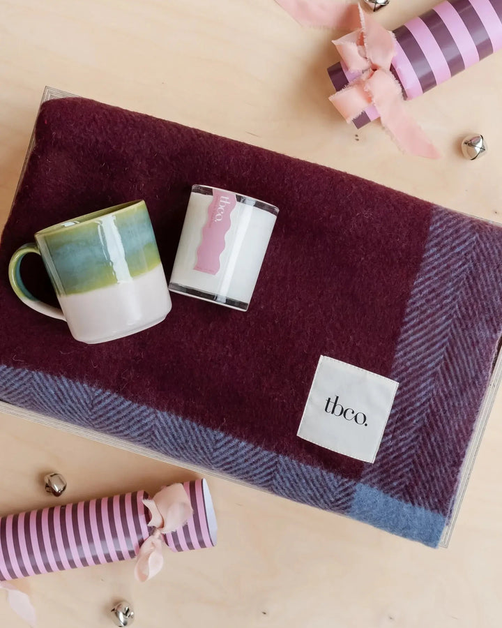 Two mugs on a plaid blanket with 'theco' branding, surrounded by striped ribbons on a wooden surface.