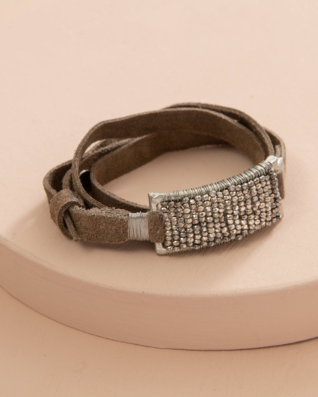 A smoky suede and silver leather wrap bracelet with a crystal embellished clasp, displayed on a beige background.