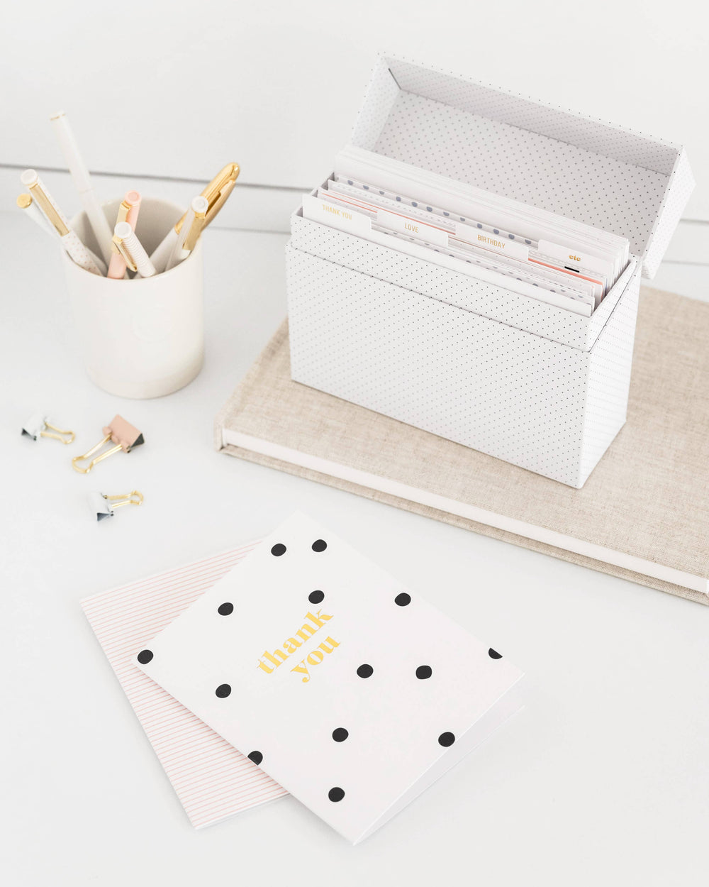 White desk with stationery items including a cup of pens, a box of cards, and a polka dot notebook.
