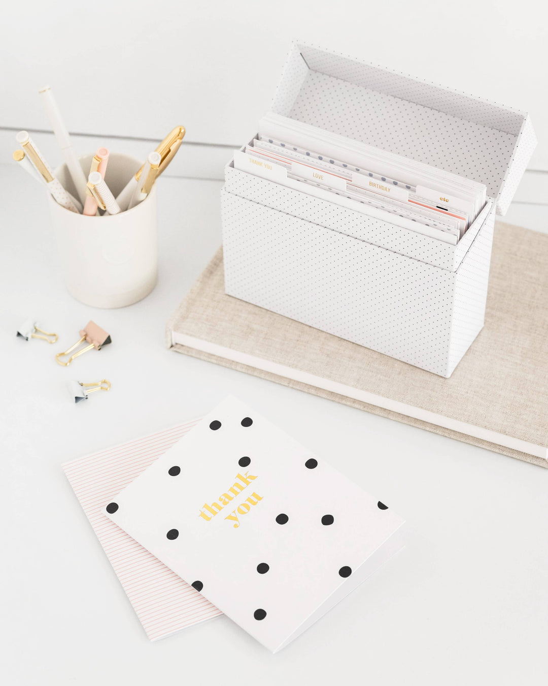 White desk with stationery items including a cup of pens, a box of cards, and a polka dot notebook.