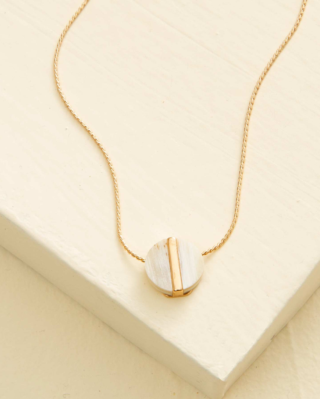 A gold-colored necklace with a circular pendant made of mixed metal and water buffalo horn, displayed on a beige background.