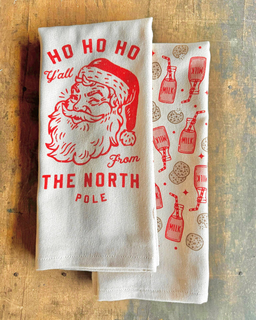 Two kitchen towels on a wooden surface, one with Santa Claus design and 'Ho Ho Ho Y'all from The North Pole', the other with milk and cookie pattern.