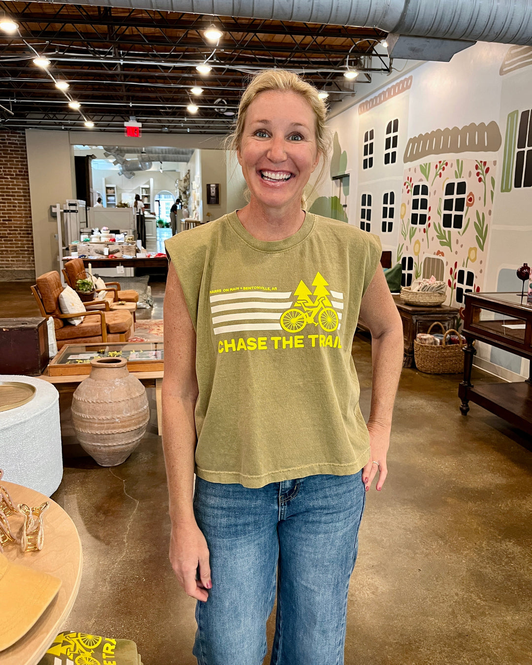A smiling blonde woman stands in a retail store interior wearing a sleeveless olive green graphic t-shirt with a yellow bicycle and 'CHASE THE TRAIL' text, paired with blue jeans.