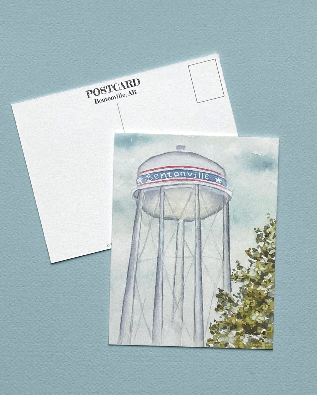 A postcard featuring a watercolor illustration of the Daytonville water tower, shown with its white back side overlapping on a textured light blue background.